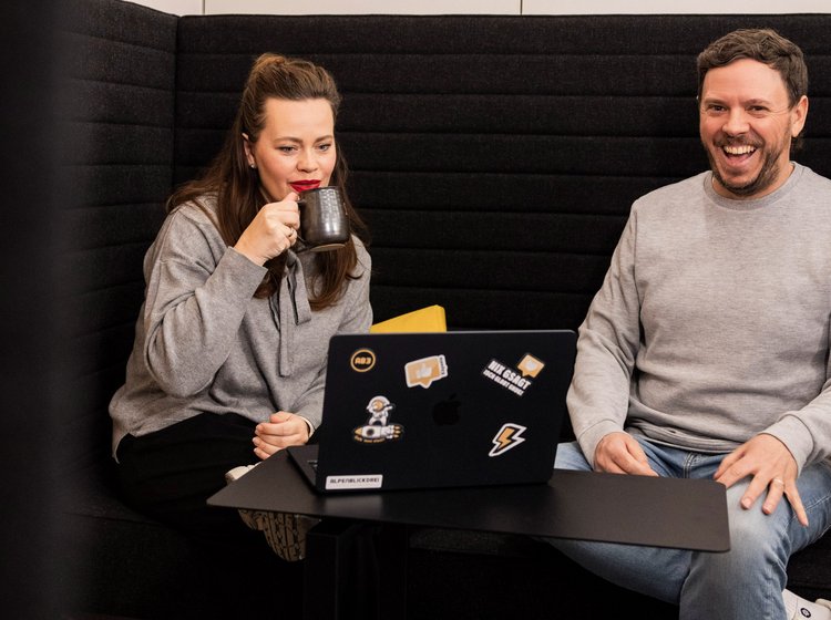 Zweiergespräch mit Kaffeepause Zwei Mitarbeitende sitzen auf einem Sofa im dunklen Raum und unterhalten sich bei Getränken.