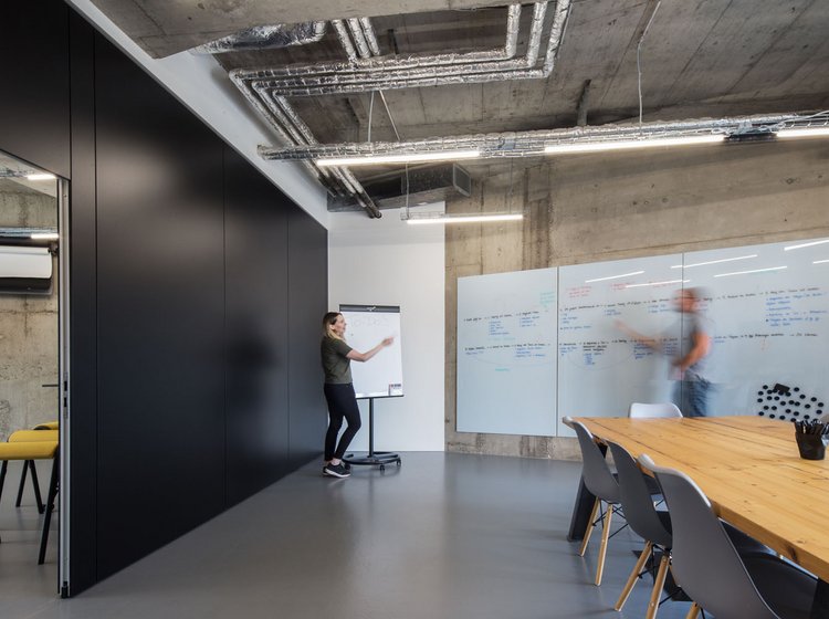 Workshopraum mit schwarzer Trennwand, Glastrennwand, Flipchart und zwei Personen in Bewegung an Whiteboards.