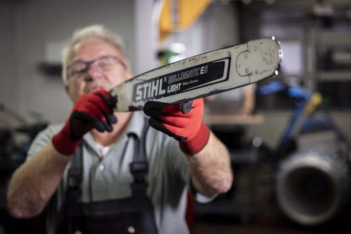 Ein Mann mit Schutzbrille und roten Handschuhen hält eine Kettensäge mit der Aufschrift „STIHL“ in die Kamera, wobei der Fokus auf der Säge liegt.