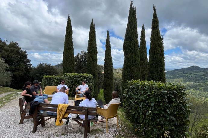 Das AB3-Team sitzt auf Gartenmöbeln unter hohen Zypressen und arbeitet im Freien, mit einer grünen Hügel-Landschaft im Hintergrund.