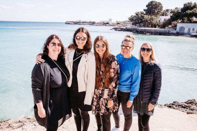Fünf Frauen stehen nebeneinander am Strand vor klarem Meerwasser, mit Küstenlinie und Häusern im Hintergrund bei sonnigem Wetter. 
