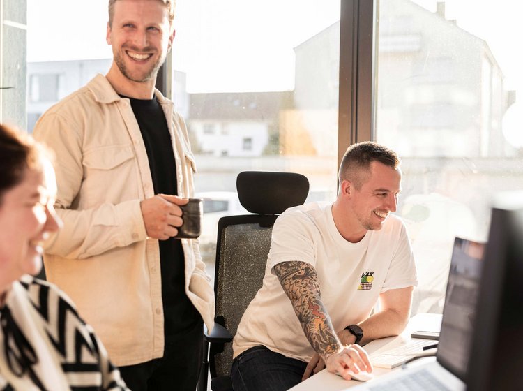 Drei Personen in einem hellen Büro lachen gemeinsam, während sie an einem Computer arbeiten und eine Tasse Kaffee trinken.