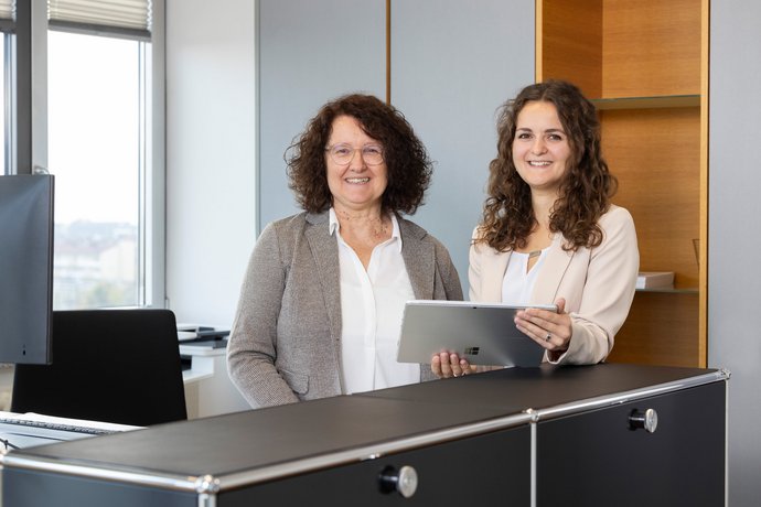 Zwei Frauen sitzen an einem Schreibtisch und lächeln in die Kamera.