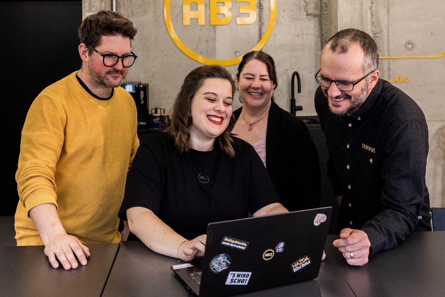 Teamrunde mit guter Stimmung Fünf Teammitglieder der Alpenblickdrei Werbeagentur lachen gemeinsam an einem Besprechungstisch im Industrie-Look.
