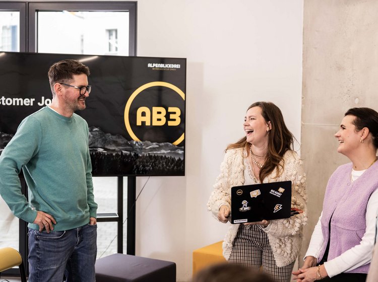 Drei Personen unterhalten sich in einem modernen Büroraum freundlich und lächelnd vor einem Bildschirm mit der Aufschrift „Customer Journey AB3“ in der Werbeagentur in Tettnang.