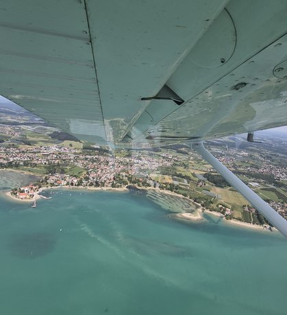 Blick aus einem Kleinflugzeug auf Wasserburg und das türkisfarbene Ufer des Bodensees.