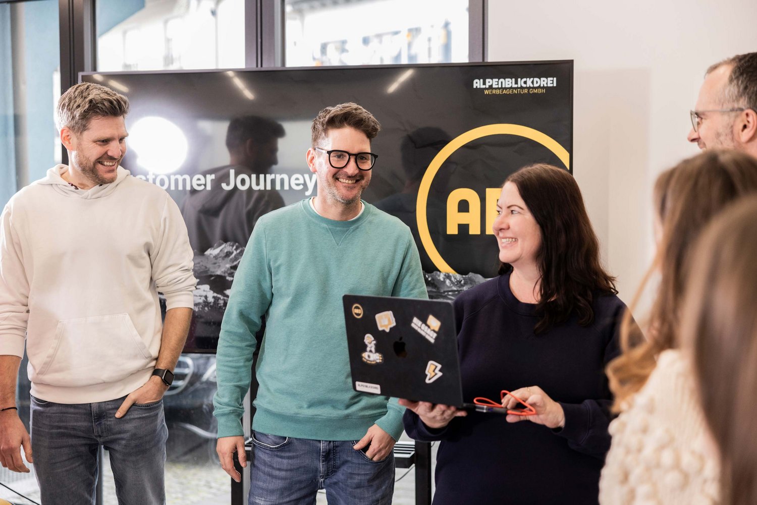 Fünf Personen stehen in einem Büro und unterhalten sich freundlich. Eine Frau hält einen Laptop und alle wirken engagiert und konzentriert bei einer Teambesprechung.