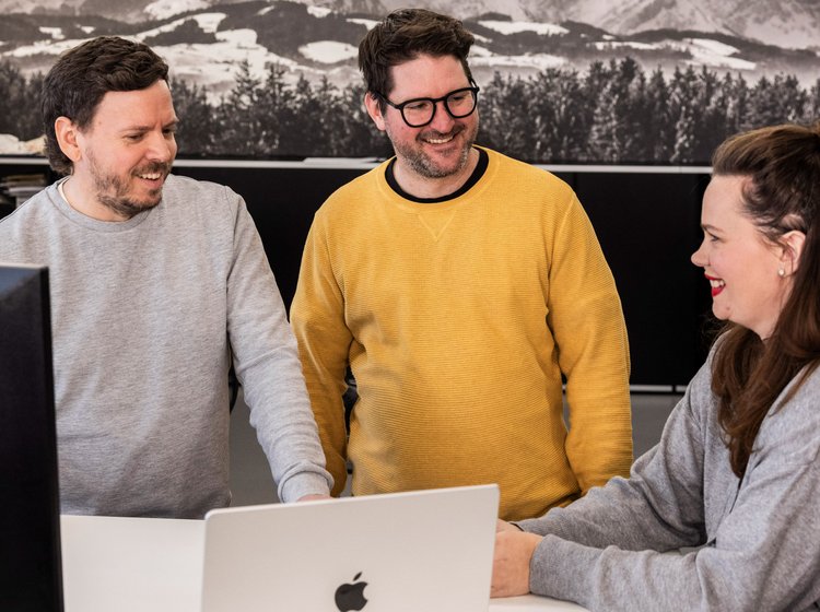 Zwei Männer im gelben Pullover stehen mit Kollegin in der modernen Teeküche der Alpenblickdrei Werbeagentur.