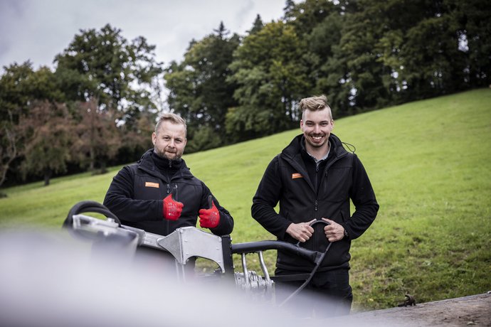 Zwei Männer in schwarzer Arbeitskleidung stehen auf einer Wiese, lächeln und geben Daumen hoch. Vor ihnen befindet sich Arbeitsgerät. Im Hintergrund Bäume und ein bewölkter Himmel.