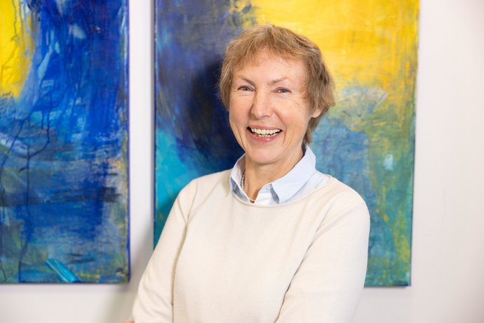 Eine ältere Frau steht vor einem Gemälde und lächelt