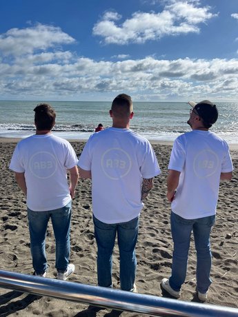 Drei Mitglieder des Alpenblickdrei-Teams blicken auf das Meer, mit weißen T-Shirts und dem AB3-Logo, vor einem klaren blauen Himmel und der Küste.