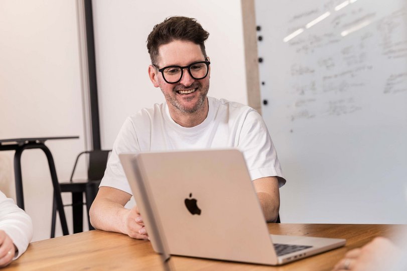 Marvin Lang sitzt an einem Tisch und arbeitet an einem Laptop. Im Hintergrund ist ein Whiteboard mit Notizen zu sehen.