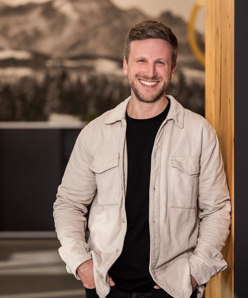 Porträt eines lächelnden Mannes mit leichter Bartstoppel, der entspannt an eine Wand gelehnt steht, trägt eine helle Jacke über schwarzem Shirt.