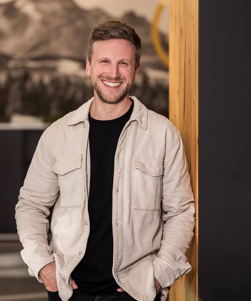 Simon Bauer Alpenblickdrei Porträt eines lächelnden Mannes mit leichter Bartstoppel, der entspannt an eine Wand gelehnt steht, trägt eine helle Jacke über schwarzem Shirt.