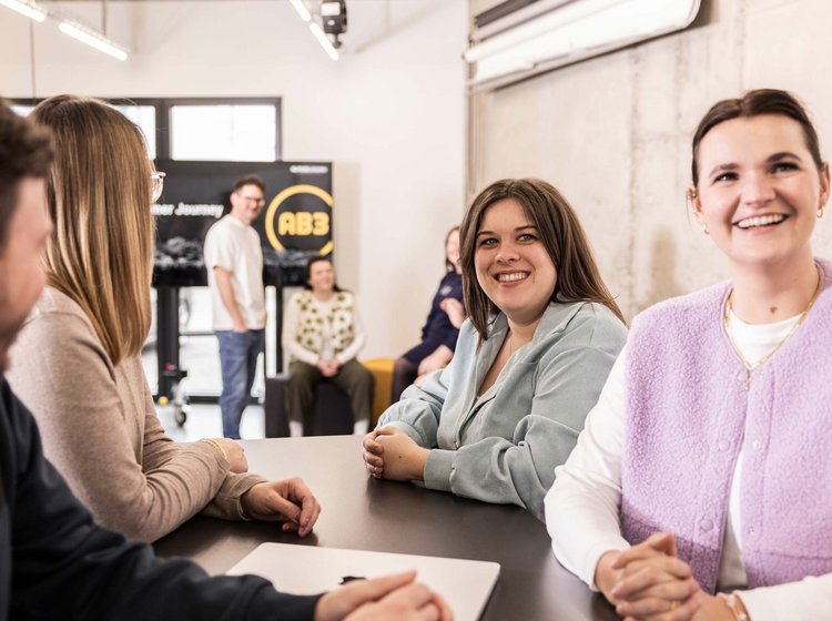 Eine Gruppe junger Menschen sitzt an einem Tisch und unterhält sich lachend in einem hellen, modernen Raum. Im Hintergrund stehen weitere Personen entspannt zusammen.