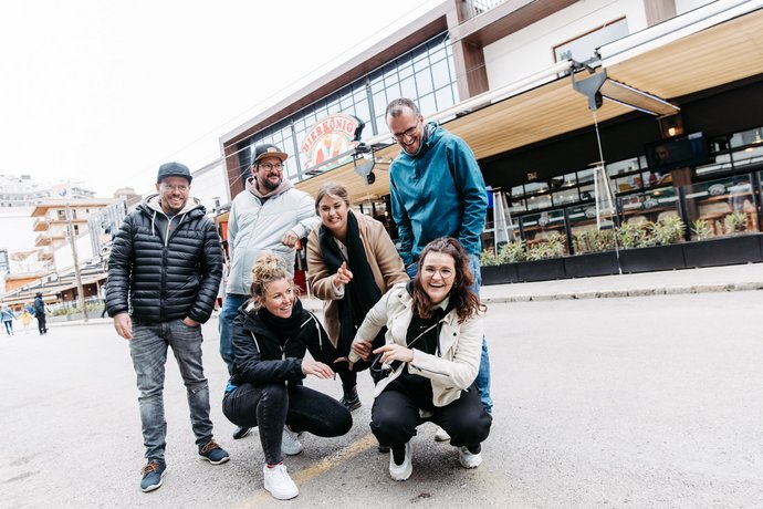 Sechs Personen posieren lachend auf einem Platz vor einem Gebäude mit Schaufenstern, entspannte Atmosphäre bei Tageslicht. 