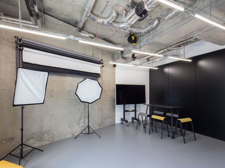 Moderner Raum mit Fotohintergrund, zwei Studiolampen, schwarzer Wand, Monitor und Konferenztisch mit gelben Stühlen unter Sichtbetondecke.