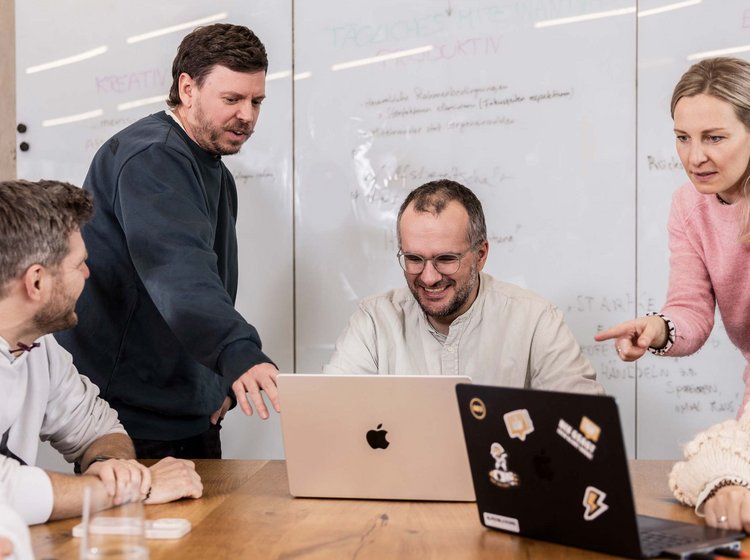 Sechs Personen sitzen und stehen um einen Tisch, zwei davon arbeiten an Laptops. Sie diskutieren gemeinschaftlich und konzentriert, vermutlich in einer Besprechung oder Teamarbeit im Büro in der Werbeagentur ALPENBLICKDREI in Tettnang. 