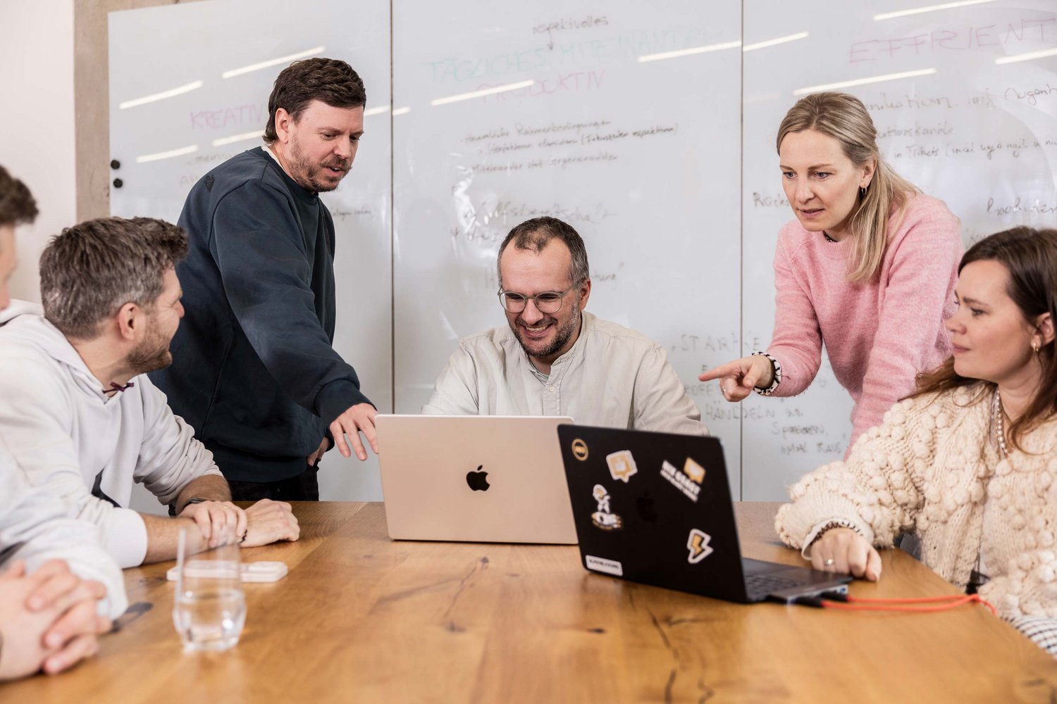 Sechs Personen sitzen und stehen um einen Tisch, zwei davon arbeiten an Laptops. Sie diskutieren gemeinschaftlich und konzentriert, vermutlich in einer Besprechung oder Teamarbeit im Büro in der Werbeagentur ALPENBLICKDREI in Tettnang. 