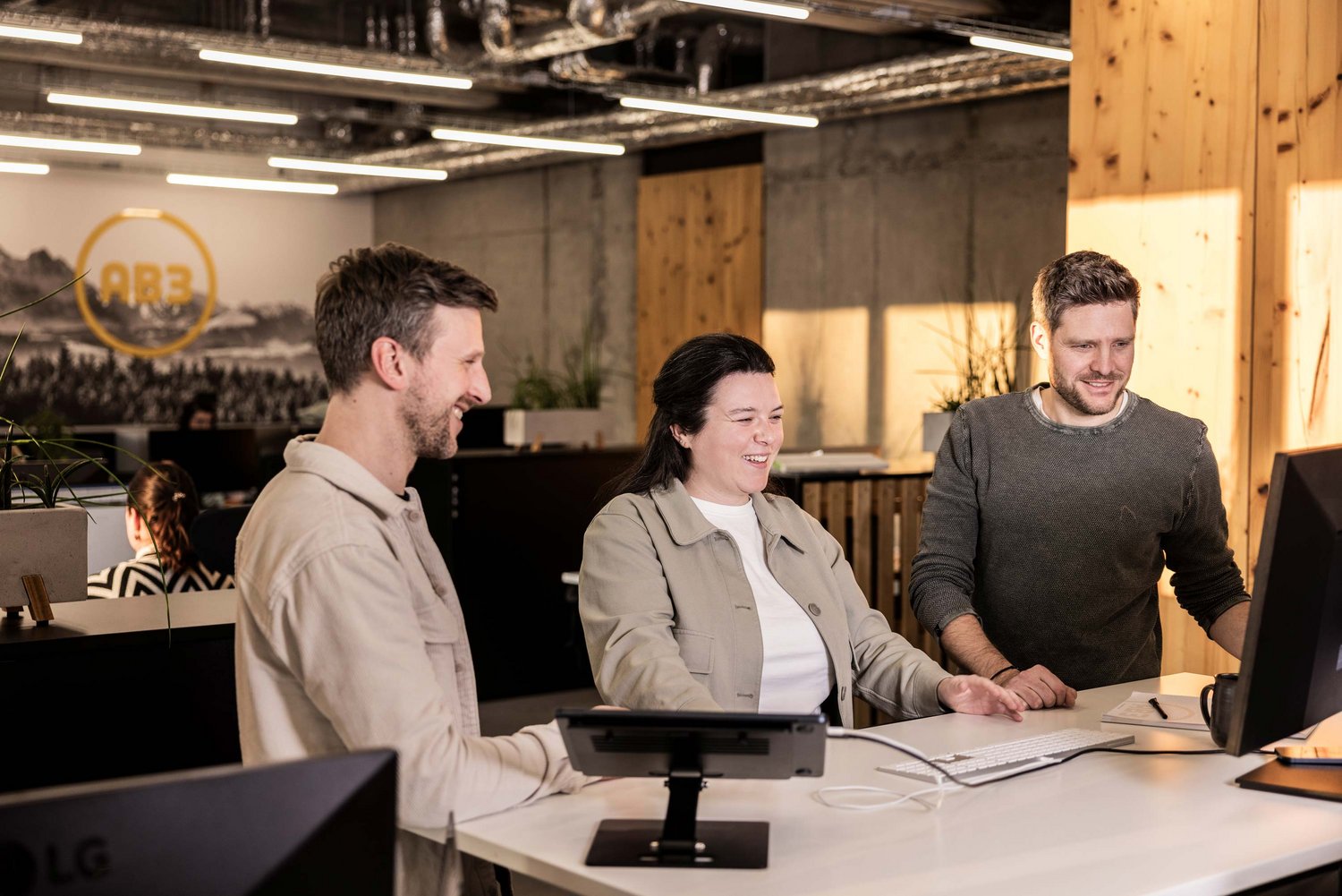 Drei Kollegen stehen gemeinsam an einem Schreibtisch und schauen lächelnd auf einen Computerbildschirm, offensichtlich in einer freundlichen Arbeitsatmosphäre in der Werbeagentur in Tettnang