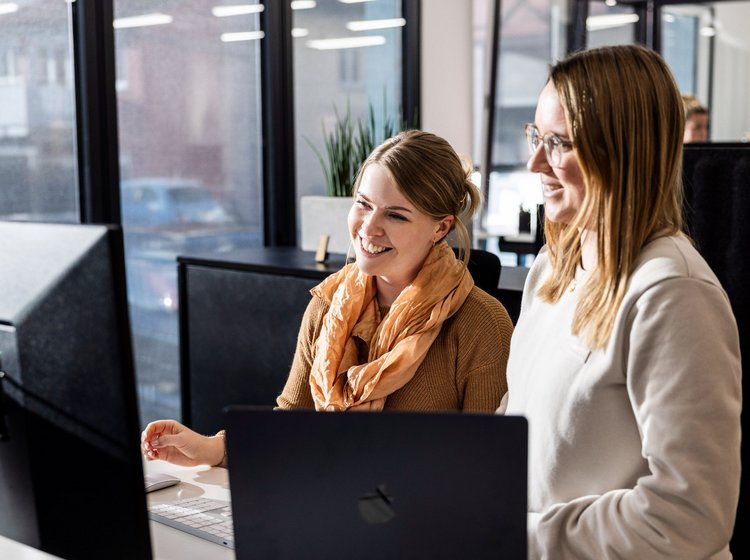 Austausch unter Kolleginnen Zwei Kolleginnen der Alpenblickdrei Werbeagentur stehen in einer modernen Büroumgebung mit Fensterfront und unterhalten sich.