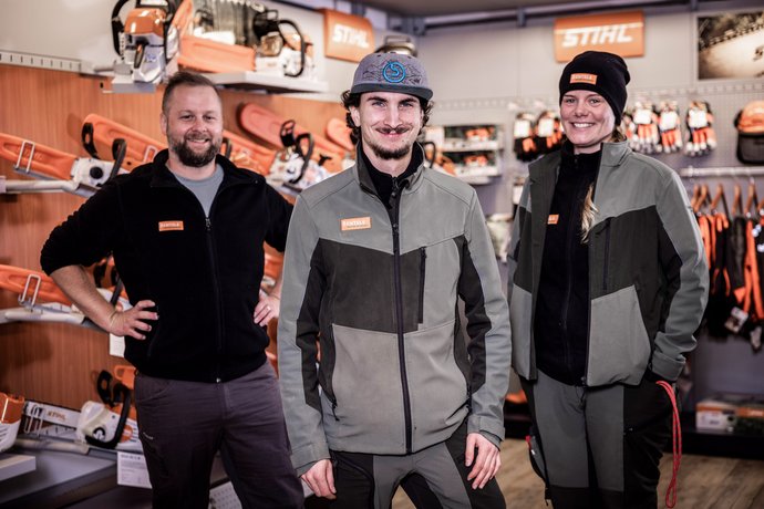 Drei Personen in Arbeitskleidung stehen lächelnd in einem Fachgeschäft für Gartengeräte, umgeben von Motorsägen und Zubehör der Marke STIHL.