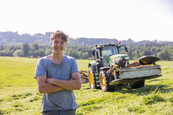Junger Landwirt vor Traktor Ein junger Landwirt steht mit verschränkten Armen vor einem Traktor auf einer Weide und lächelt in die Kamera.