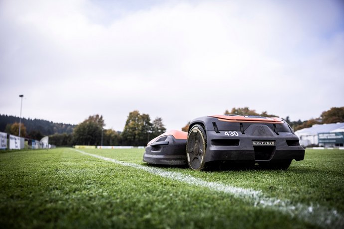 Zwei autonome Rasenmähroboter fahren auf einem gepflegten Fußballfeld nebeneinander entlang einer weißen Spielfeldlinie.