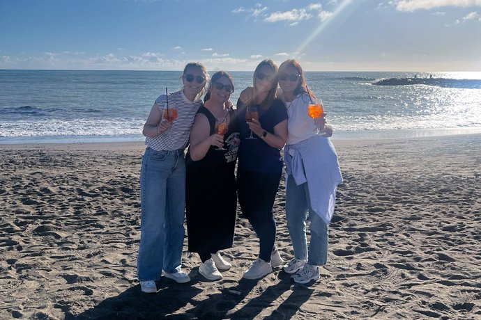 Vier Mitglieder des Alpenblickdrei-Teams stoßen mit Aperol Spritz an, lachen und genießen das sonnige Wetter am Strand, mit dem Meer im Hintergrund.