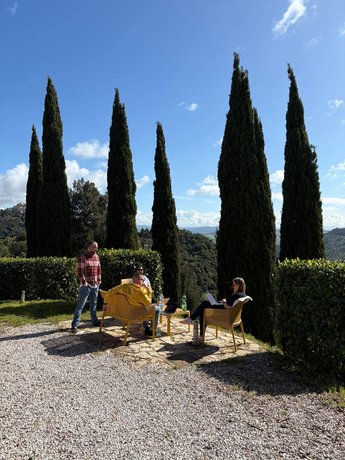 Ein Teil des AB3-Teams arbeitet entspannt unter Zypressen im Freien, mit Laptops auf einem Steintisch, vor einer beeindruckenden Aussicht auf die toskanische Landschaft.