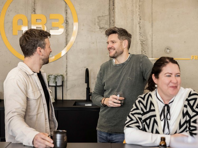 Drei Personen unterhalten sich locker in einem modernen Büro mit Betonwänden, zwei Männer stehen, eine Frau sitzt am Tresen.