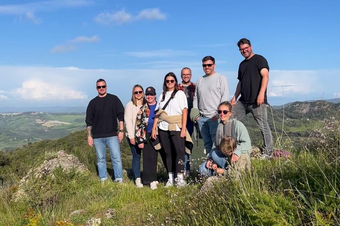 Ein Teil des AB3-Teams posiert auf einem Hügel mit herrlichem Blick auf die toskanische Landschaft, mit sonnigem Wetter und grüner Umgebung. Alle lächeln in die Kamera.