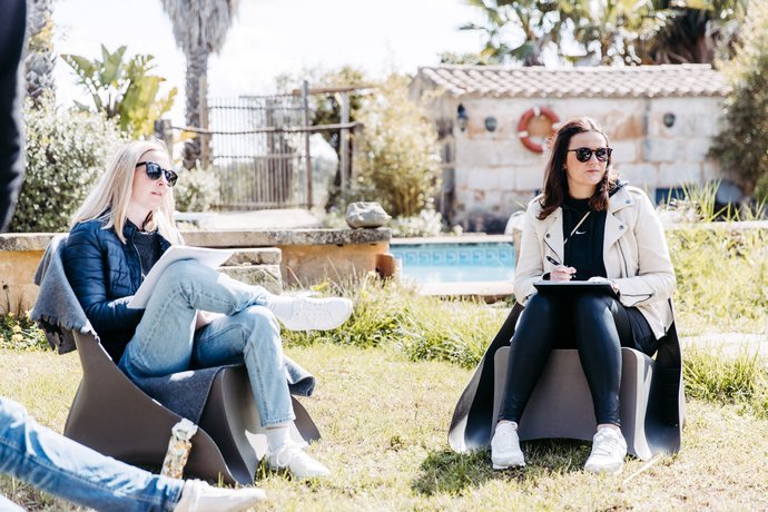 Zwei Frauen sitzen auf modernen Outdoor-Stühlen auf einer Rasenfläche mit Pool und mallorquinischem Haus im Hintergrund, bei sonnigem Wetter.