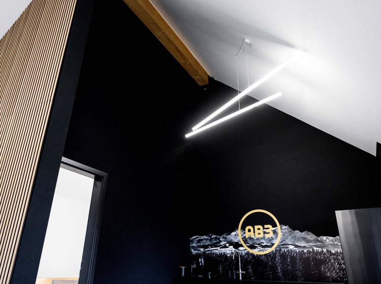 Innenaufnahme eines Raums mit schwarzer Wand, an der ein großes Bild von Bergen und das gelbe Logo „AB3“ zu sehen sind. Die Decke ist weiß mit hölzernem Balken, daran hängen zwei moderne, lange LED-Lichtleisten. Eine helle Türöffnung führt in einen angrenzenden Raum.