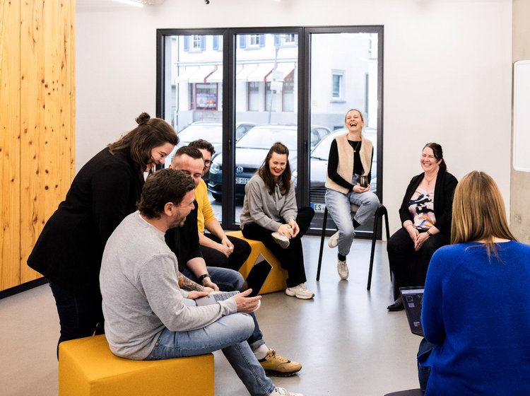 Meeting im hellen Büroraum Mehrere Mitarbeitende der Alpenblickdrei Werbeagentur in lockerer Runde mit Sofas und Sesseln im offenen Büro.