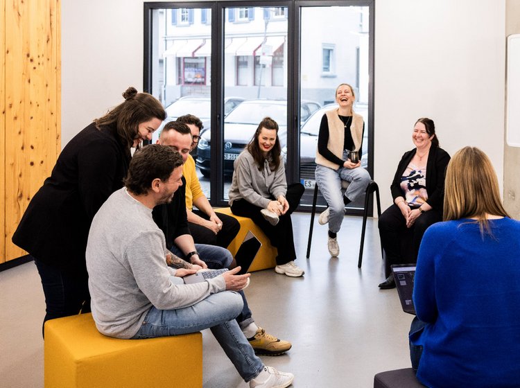 Mehrere Mitarbeitende der Alpenblickdrei Werbeagentur in lockerer Runde mit Sofas und Sesseln im offenen Büro.