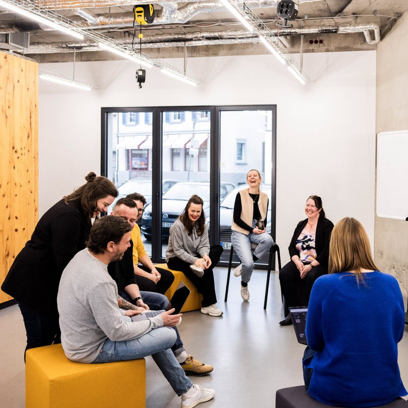 Mehrere Mitarbeitende der Alpenblickdrei Werbeagentur in lockerer Runde mit Sofas und Sesseln im offenen Büro.