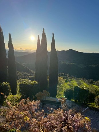 Ausblick aus unserer Unterkunft I Casali Toscana, Blick auf einen idyllischen Garten mit Zypressen und Sitzgelegenheiten, vor einer hügeligen Landschaft im Sonnenuntergang.