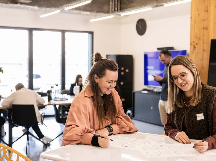 Zwei Frauen arbeiten gemeinsam an einem Projektplan oder einer Zeichnung an einem Tisch in einem modernen, hellen Büro. Im Hintergrund sind weitere Personen in konzentrierter Arbeitsatmosphäre zu sehen.