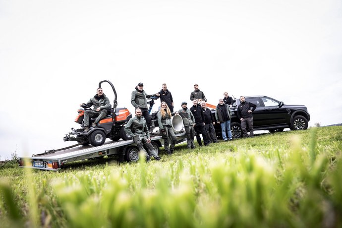 Eine Gruppe von elf Menschen steht und sitzt auf einem Anhänger, auf dem ein Rasenmäher sowie weitere Ausrüstungsgegenstände geladen sind, neben einem schwarzen Auto auf einer Wiese. Die Szene wirkt professionell und gemeinschaftlich.