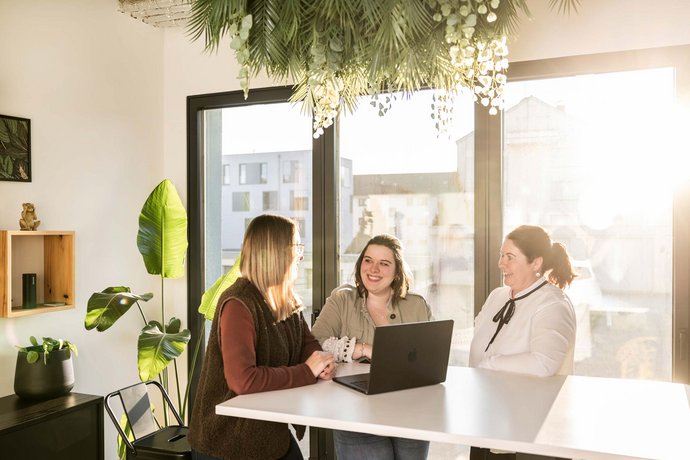 Drei Frauen sitzen gemeinsam an einem Tisch mit Laptop und unterhalten sich freundlich in einem hellen, modernen AB3 Büro mit großen Fenstern und Pflanzen in Tettnang.