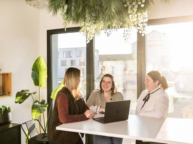 Drei Frauen sitzen gemeinsam an einem Tisch mit Laptop und unterhalten sich freundlich in einem hellen, modernen AB3 Büro mit großen Fenstern und Pflanzen in Tettnang.