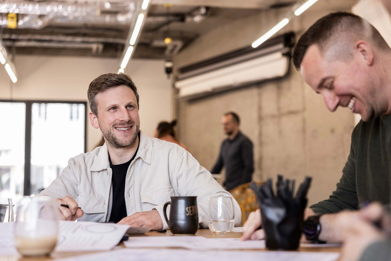 Zwei Männer sitzen an einem Tisch, lächeln und arbeiten gemeinsam an Unterlagen, sie wirken entspannt und konzentriert in einem modernen Büro.