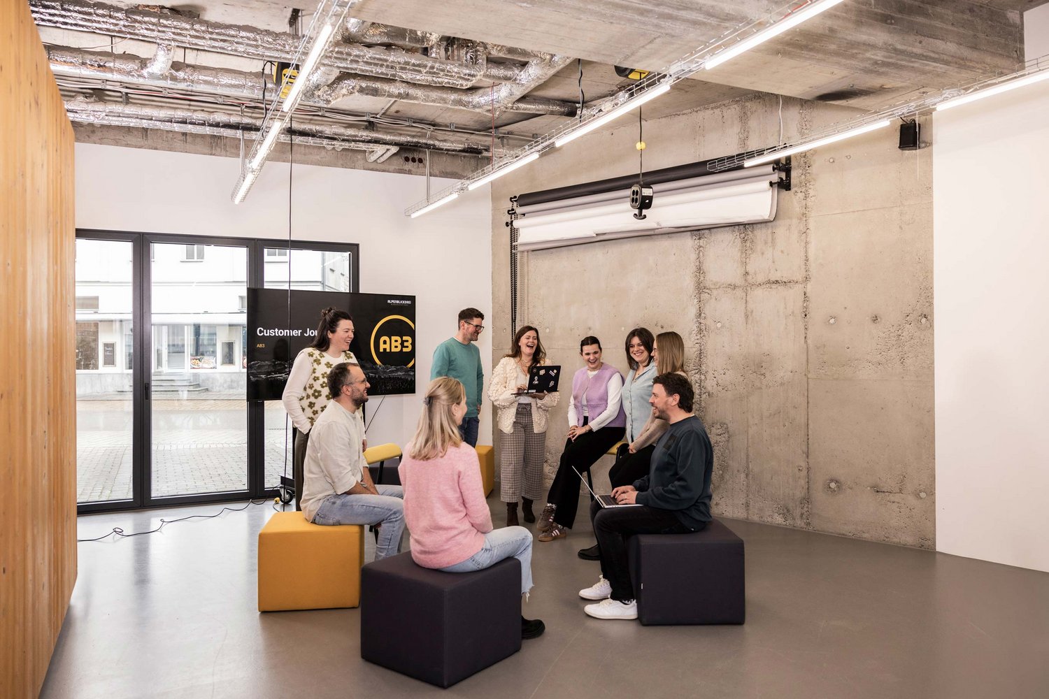 Eine Gruppe von neun Personen sitzt und steht in einem modernen, offenen Besprechungsraum und führt eine informelle Teamdiskussion.