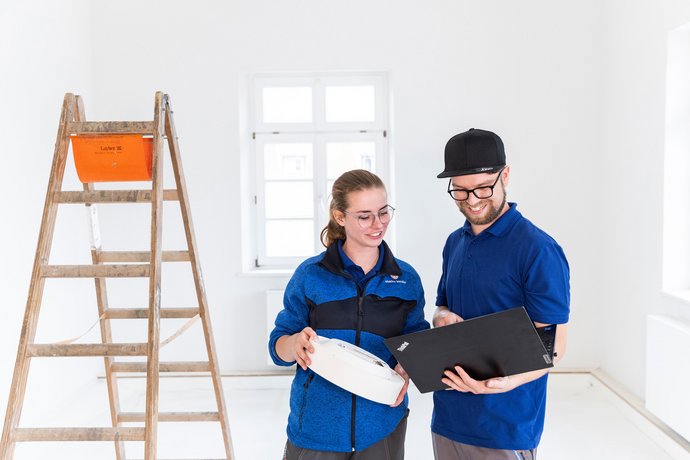 Elektro Kessler – Licht & Beleuchtung Zwei Elektriker in blauer Arbeitskleidung besprechen Unterlagen vor einer Leiter im hellen Raum.