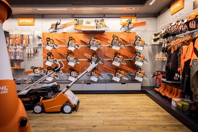Ladenwand mit verschiedenen Benzin-Kettensägen und Aufsätzen der Marke Stihl, dazu passende Schnittschutzkleidung und Zubehör wie Helme und Schutzstiefel, alles in orange und schwarz gehalten. Ein Rasenmäher steht im Vordergrund auf dem Holzboden.