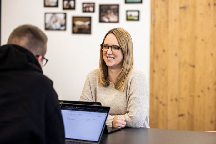 Teamgespräch im Büro Zwei Personen im Gespräch an einem Schreibtisch, im Hintergrund eine Bilderwand und Holzwand.