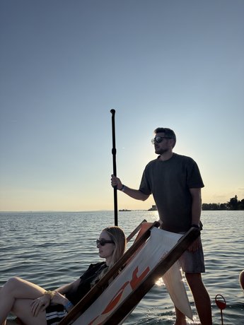 Zwei Personen entspannen auf einer schwimmenden Plattform, während ein Teammitglied über den Bodensee paddelt.