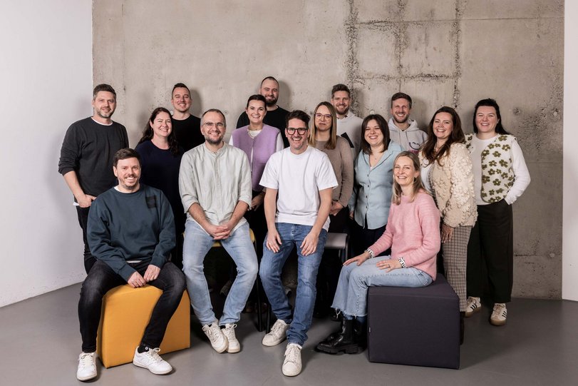 Gruppenbild von 15 Personen unterschiedlichen Alters, die lächelnd vor einer grauen Betonwand stehen und sitzen, entspannte und freundliche Stimmung.