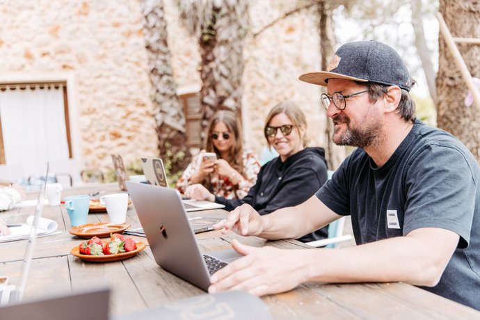 Mann mit Cap arbeitet an Laptop an einem Holztisch im Freien, daneben Snacks und Getränke, weitere Personen im Hintergrund vor Steinmauer. 